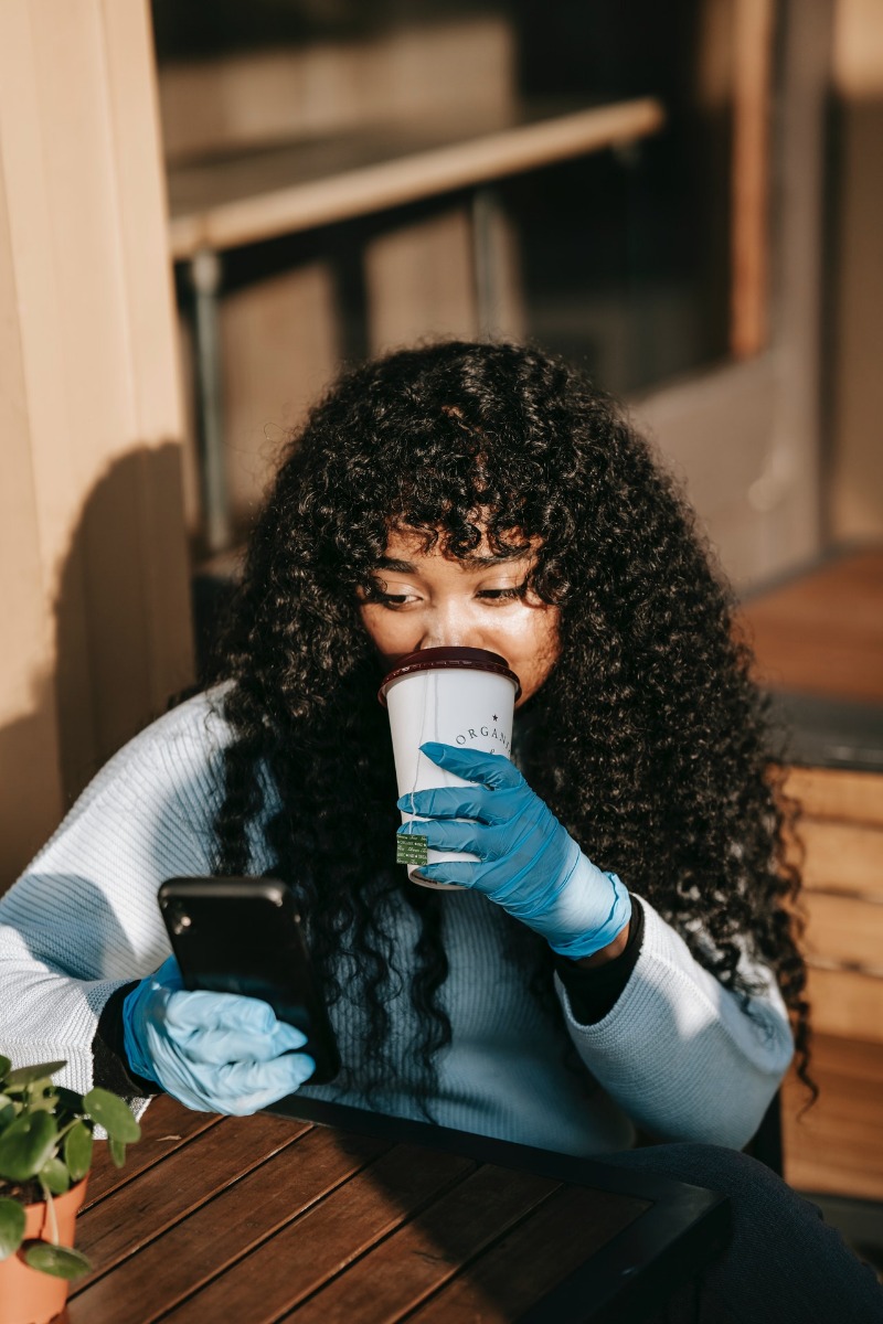 curly haired woman sipping on a hot drink while looking at her smart phone